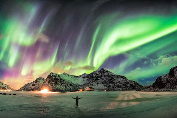 Man standing with arms upraised in the snow, under a vibrant green and purple Aurora Borealis