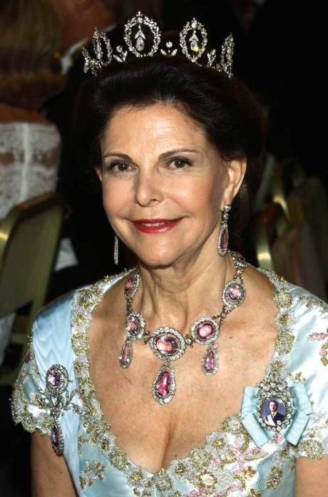 Queen wearing a diamond crown and fancy dress, around her neck is a necklace of large pink Imperial Topaz haloed by diamonds