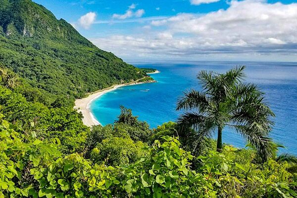 Aerial view of the Dominican Republic, green jungle mountain on one side, vivid blue ocean on the other, with a thin crescent of white sandy beach