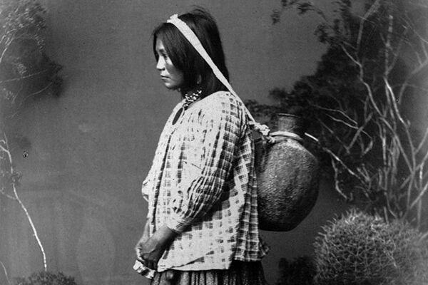 Black and white photo of an Apache woman carrying water on her back