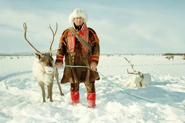 Sami man in traditional fur outfit with white reindeer, standing in a snowy field.